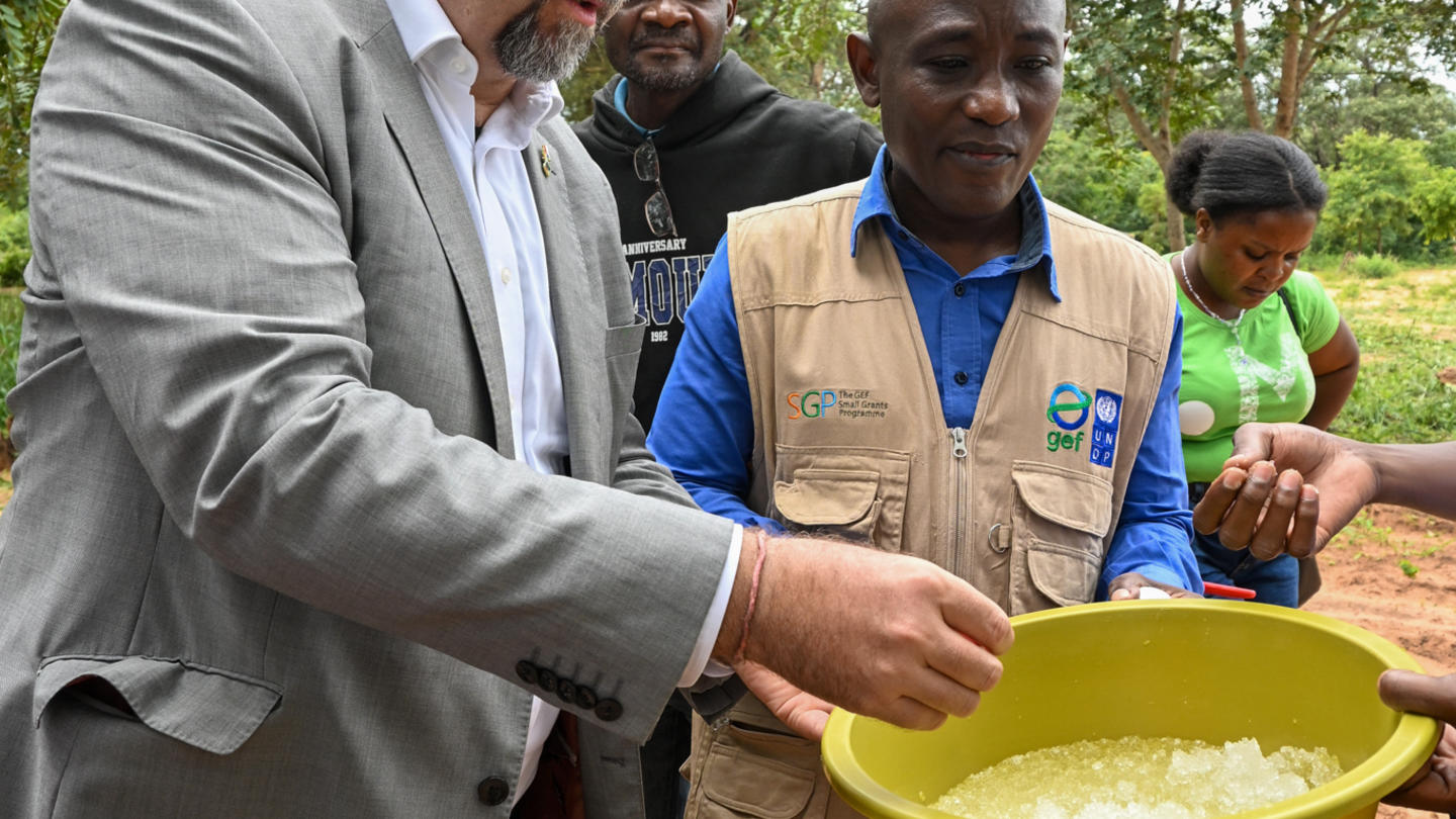 A man in a gray suit hands a yellow bowl to a person wearing a blue vest on a dirt field.