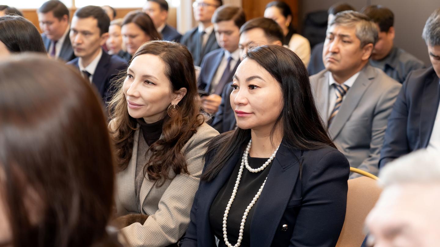 Audience seated in a conference room; foreground woman with dark hair and pearl necklace.