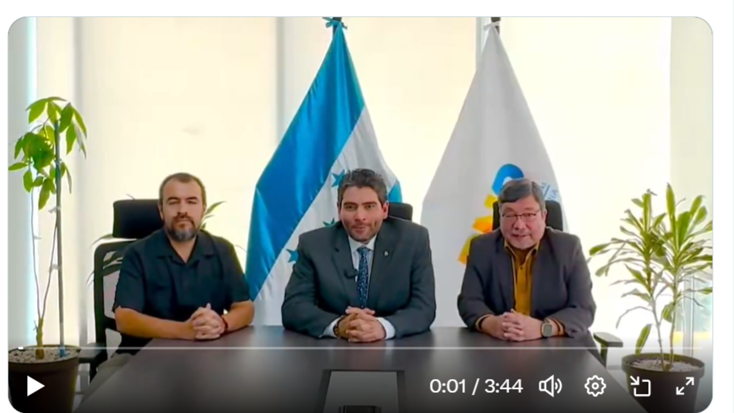 Video shows three men in suits sitting at a panel with microphones, flags behind them.