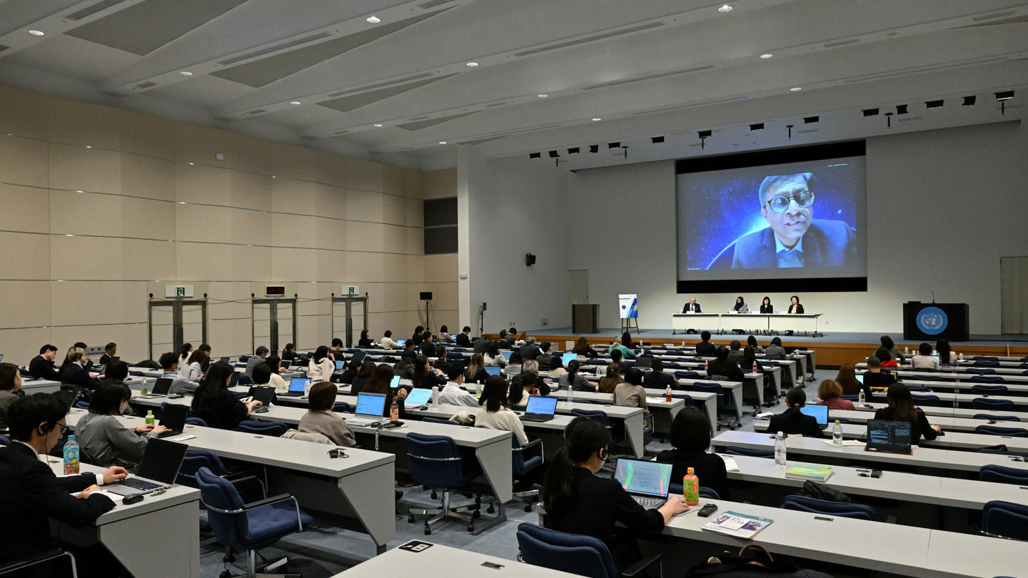 Photograph of a large lecture hall with attendees facing a podium; a big screen shows a speaker.