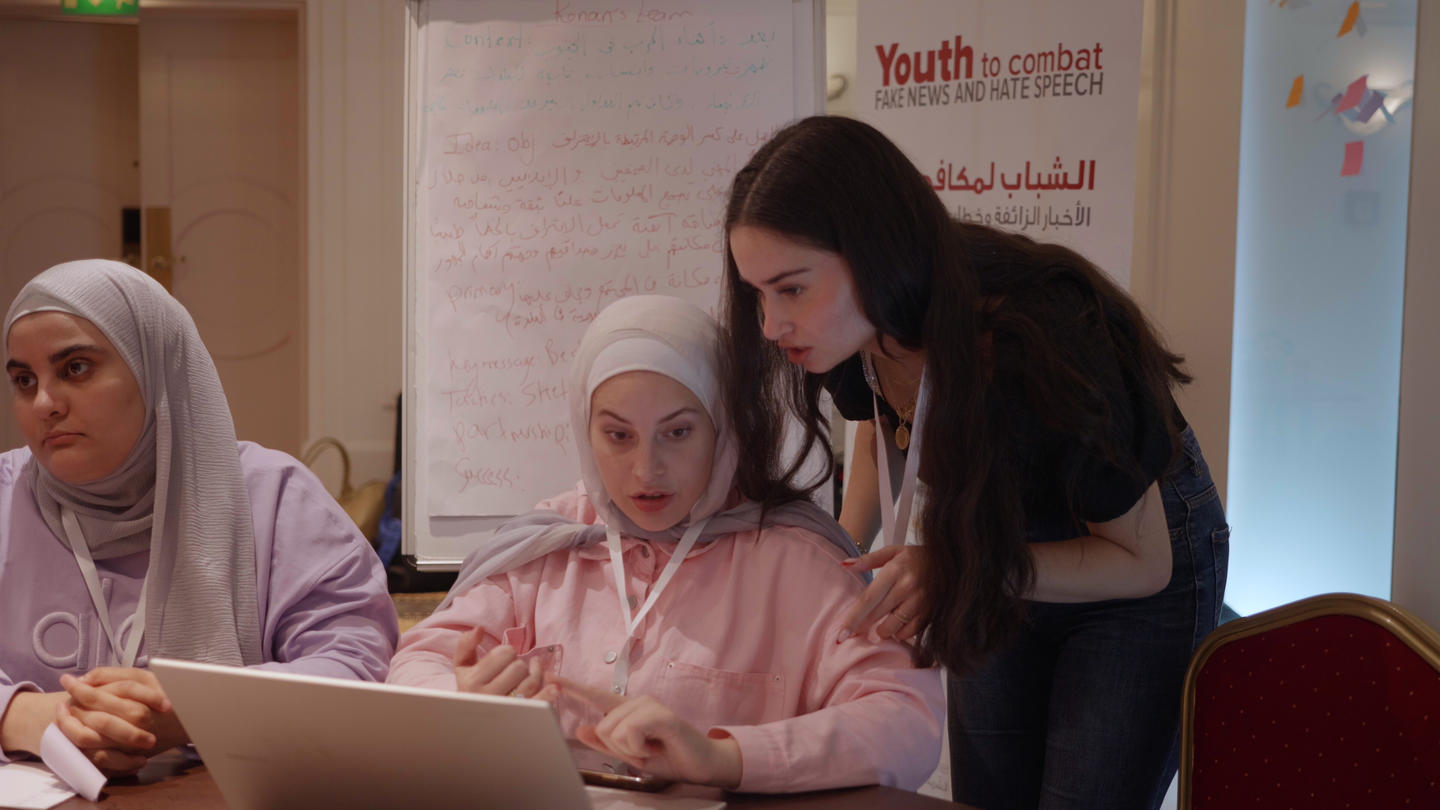 Three students at a table with laptops; a standing mentor leans over to help.