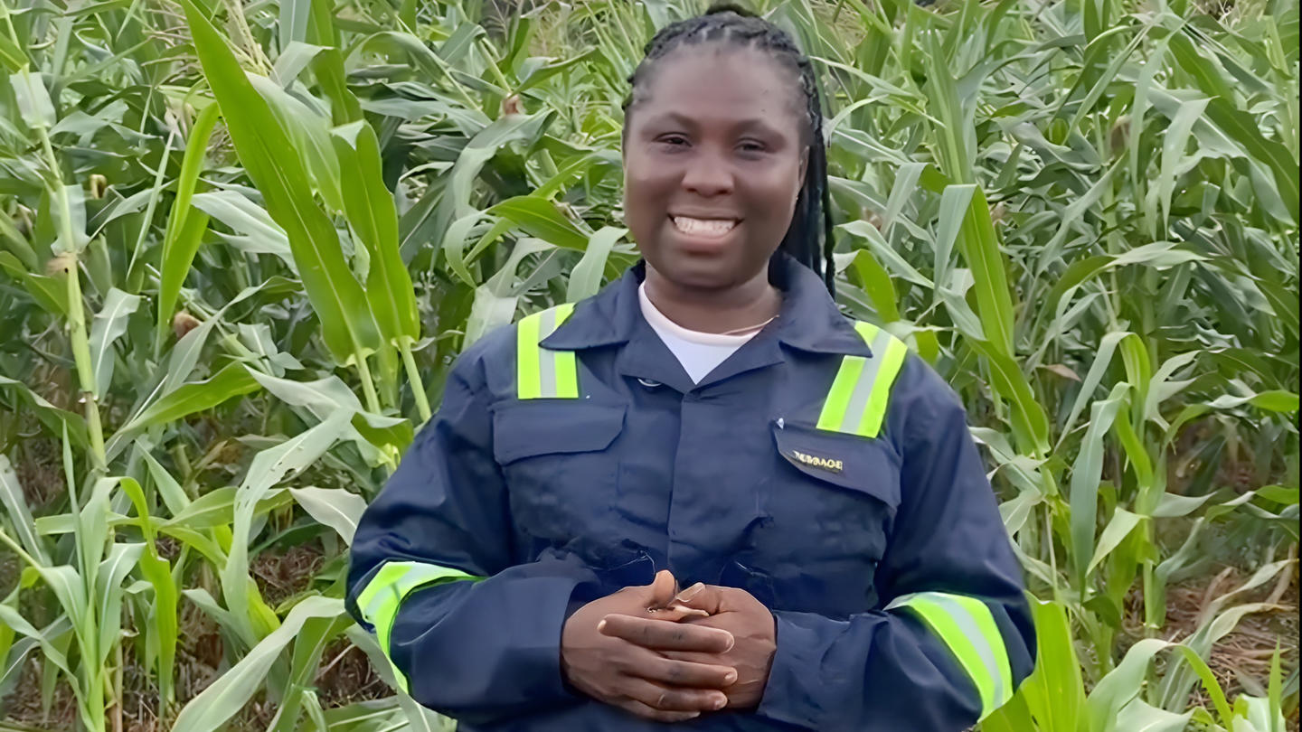 Photograph of a worker in safety uniform with reflective stripes in a cornfield, hands clasped.