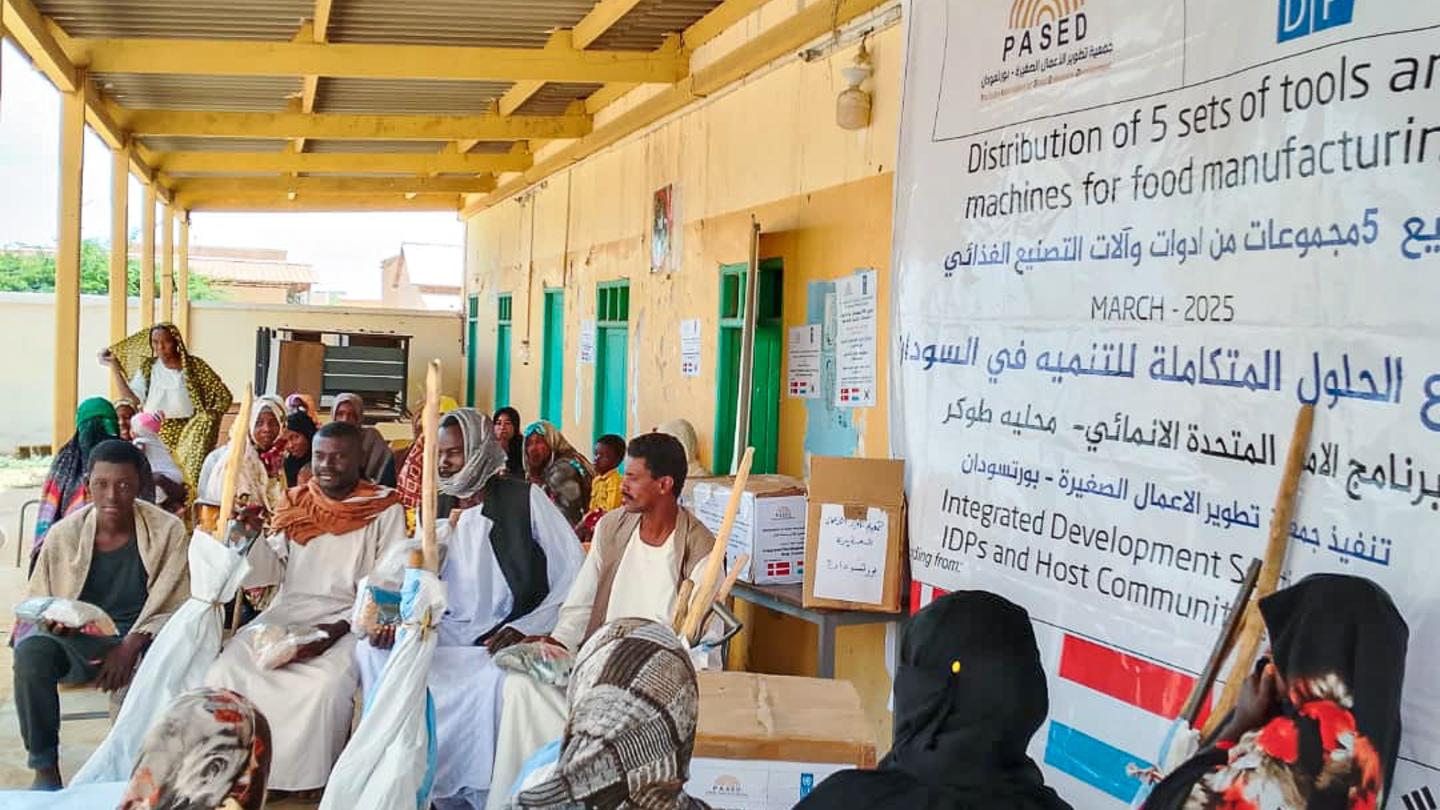 Women in headscarves sit with others under a wooden shelter; a banner with logos on the wall.