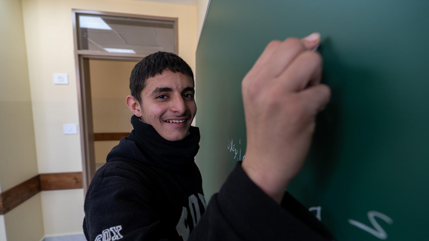 Person in a black hoodie writes on a green chalkboard in a hallway.