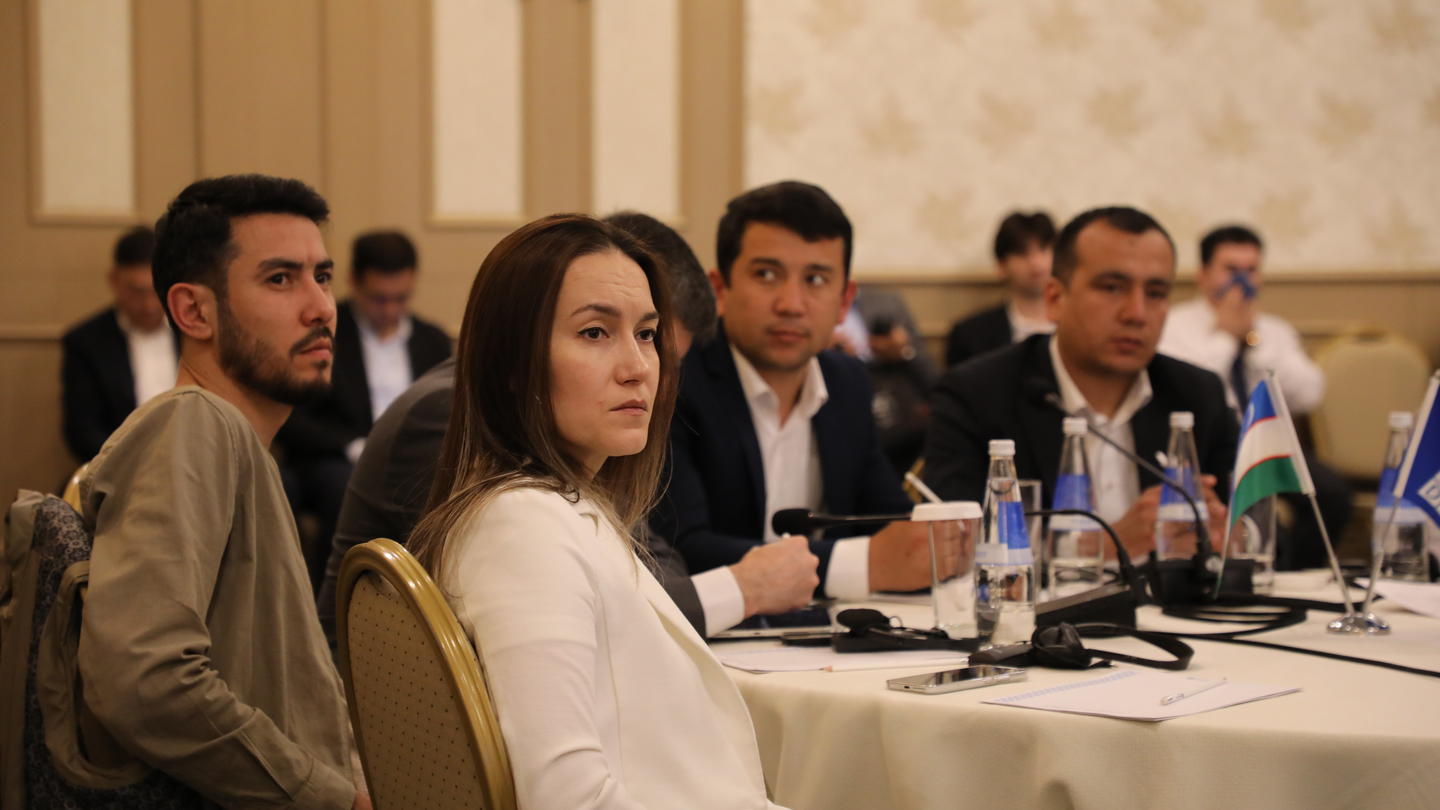 Group of professionals at a formal conference table in a meeting room; foreground woman blurred.