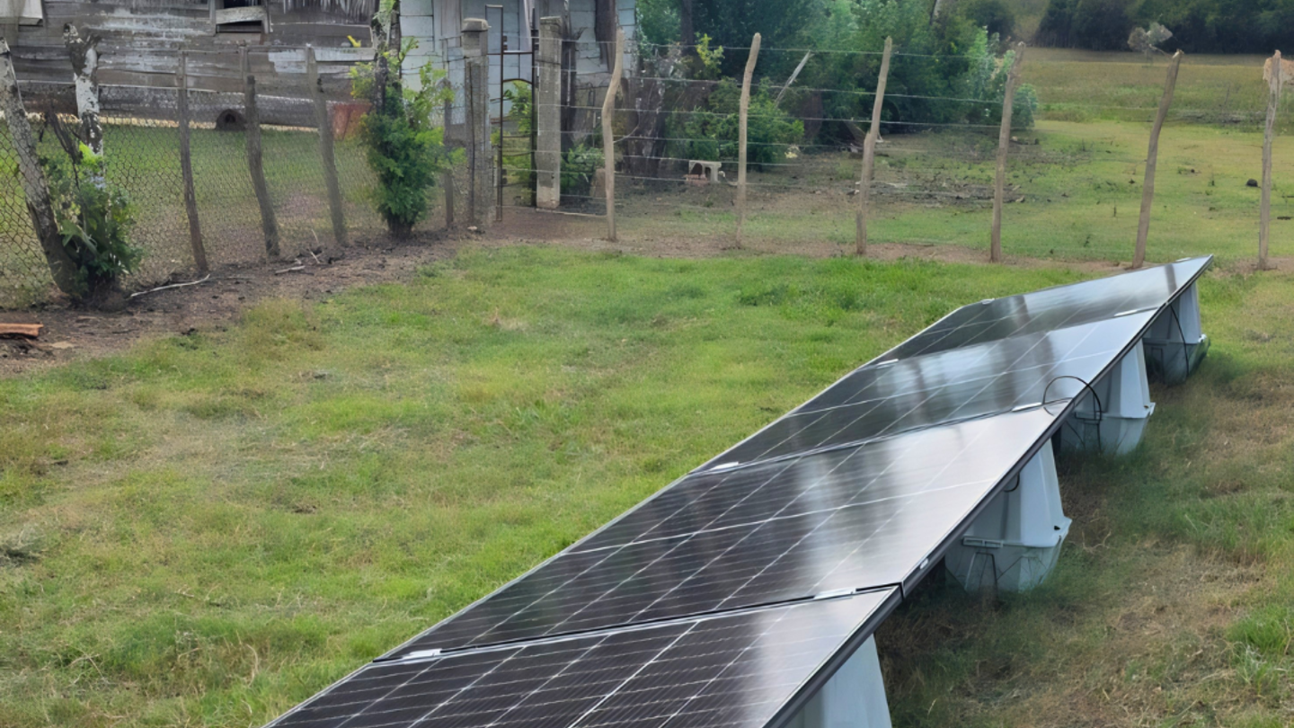 Long row of solar panels on a grassy field beside a thatched hut and fence.
