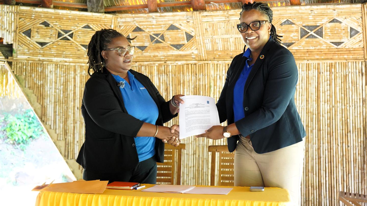 Two people exchange a document at a ceremony, standing behind a yellow table.