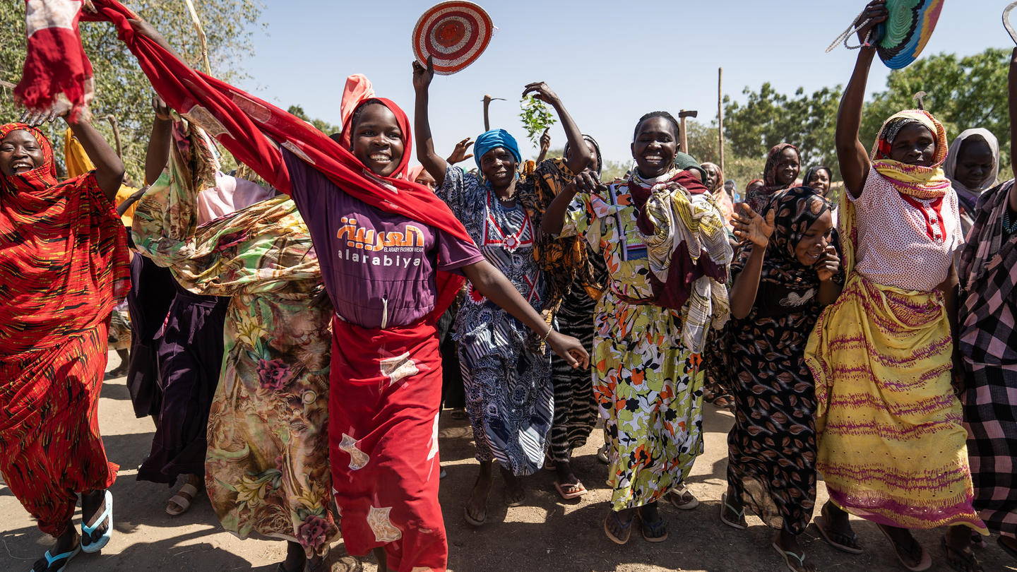 Group of women in bright traditional dresses dancing outdoors, raising arms with round fans.