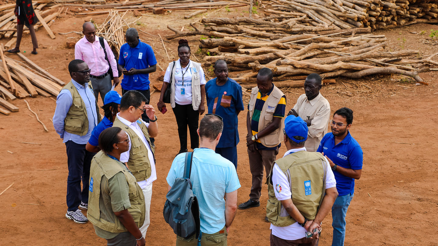 Group of people including UN partners standing in a circle on red dirt with stacked logs behind.