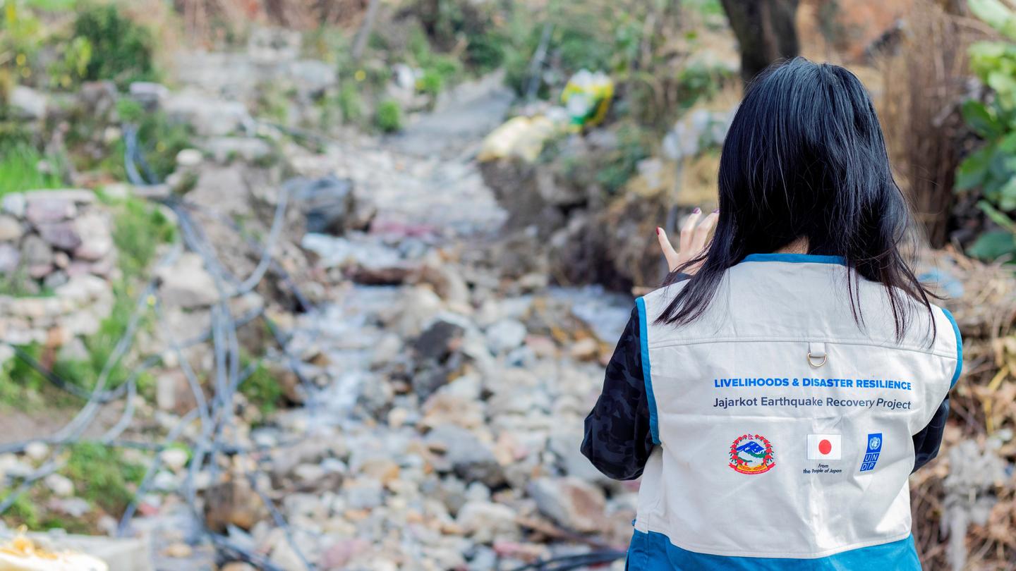 Back view of a person wearing a gray vest with logos, standing beside a rocky stream.