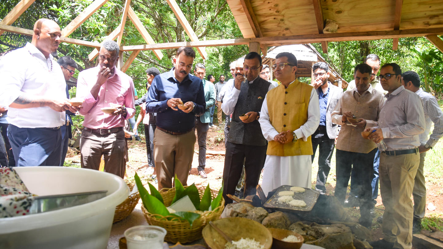 Group of people outdoors under a wooden pavilion, gathered around a table of food.