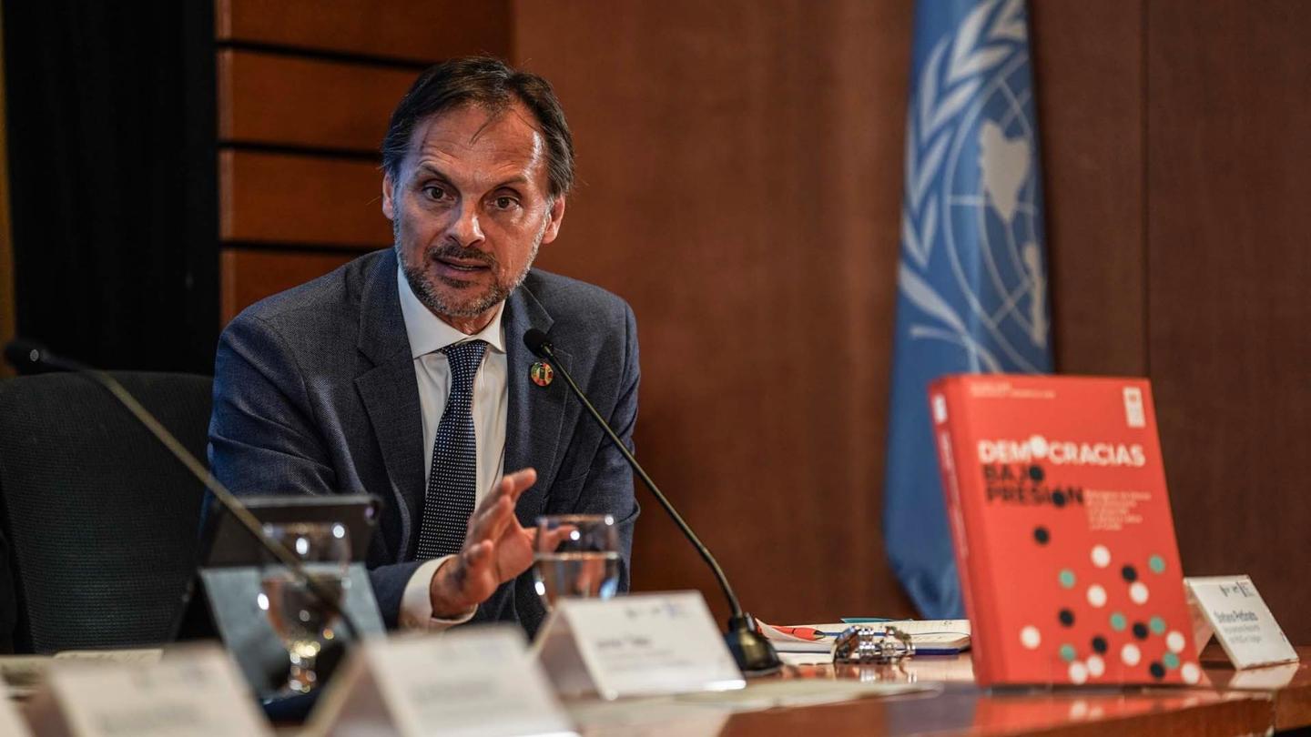 Photograph of a suited man at a conference table with microphones, UN flag in the background.