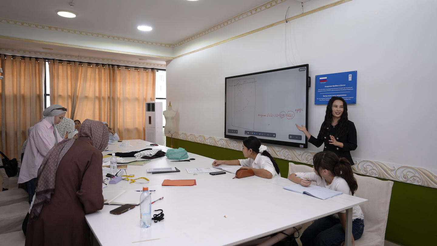 People wearing hijabs around a long table in a conference room; presenter at front with screen.
