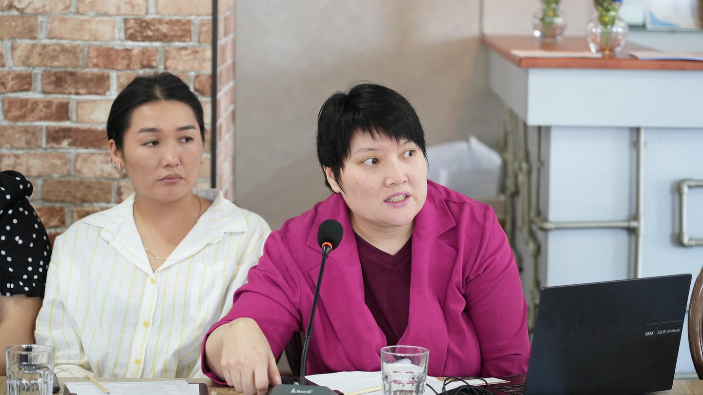 Two women sit at a table in a meeting, looking over papers with laptops nearby.