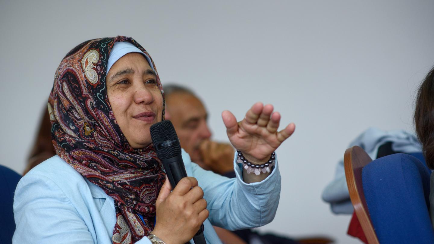 Woman in a patterned hijab speaks into a handheld microphone at a conference.