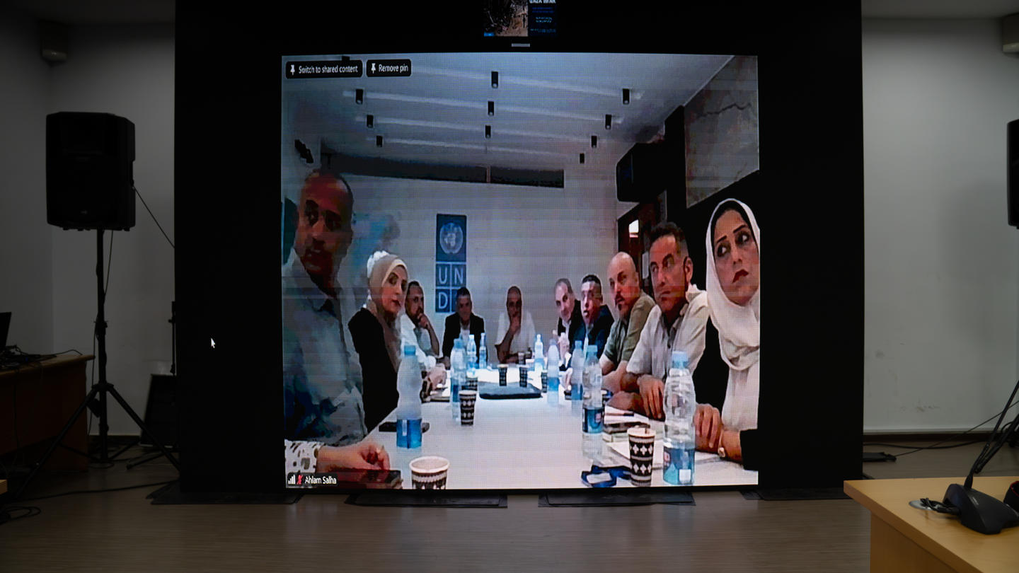 A group of people around a long conference table is shown on a large video screen in a dark room.