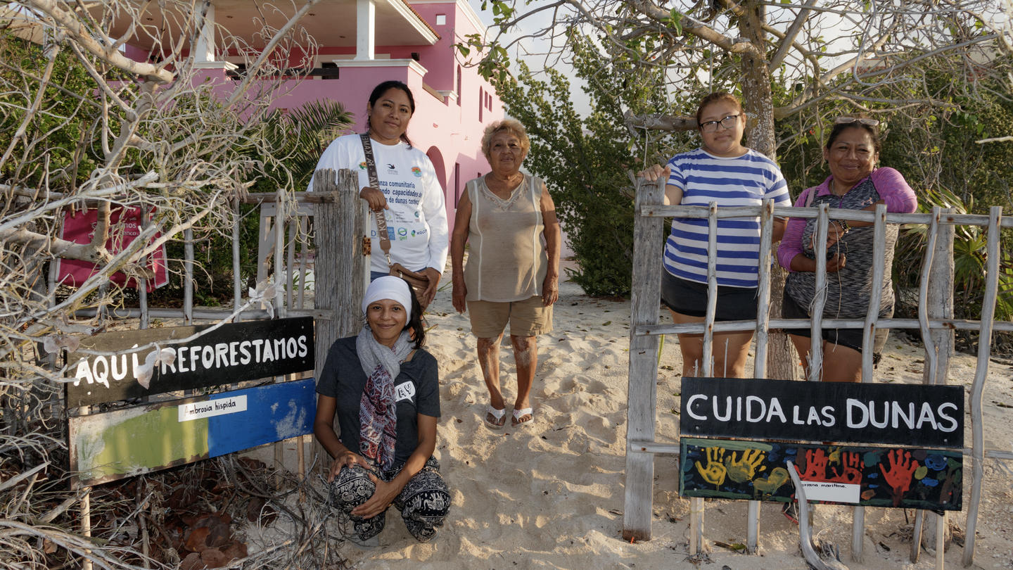 Group of people on a sandy dune path beside signs: CUIDA LAS DUNAS, AQUI FORESTACION