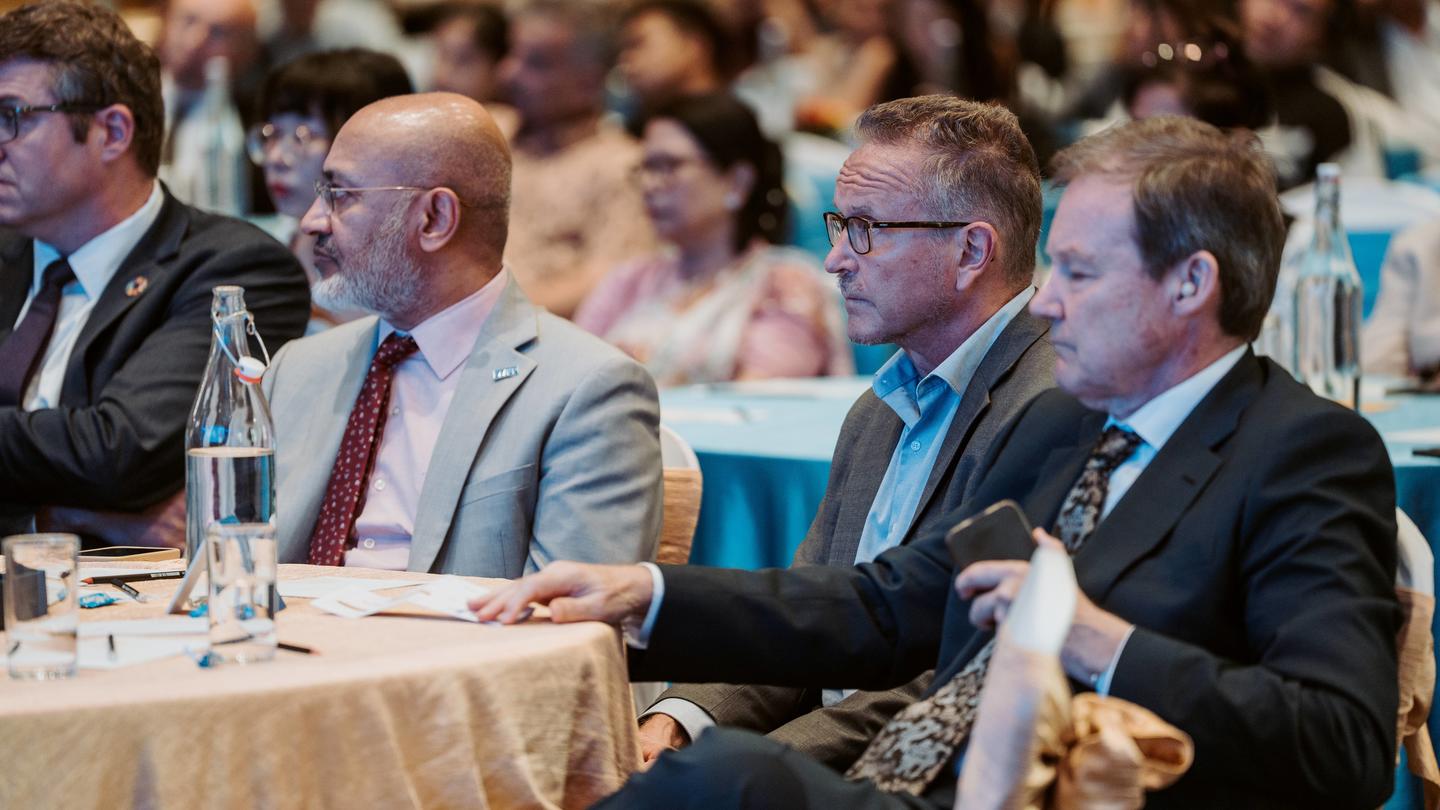 Photo: three men in suits seated at a round table at a conference, crowd in background.