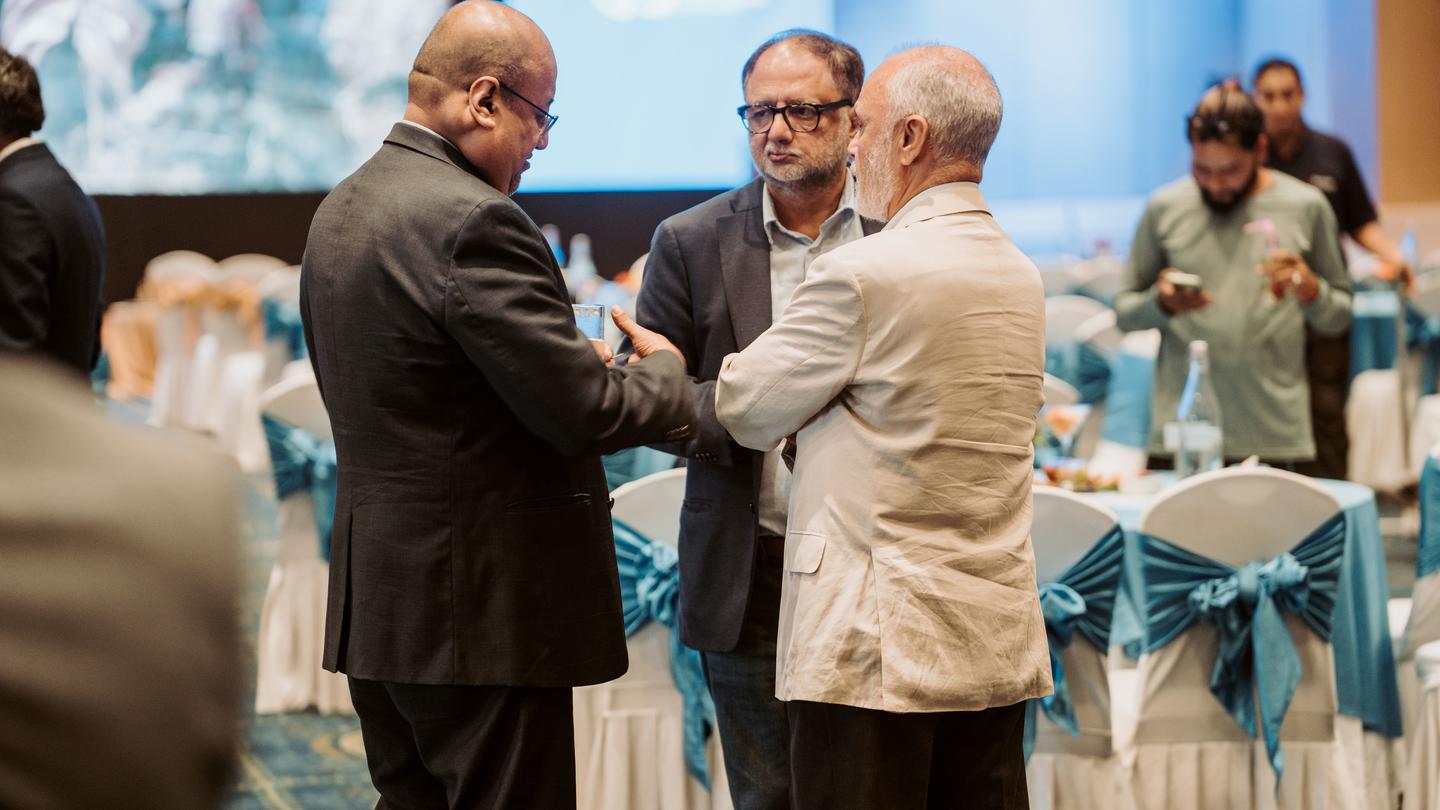 Photo shows two men in suits shaking hands at a formal event, teal banquet tables in background.