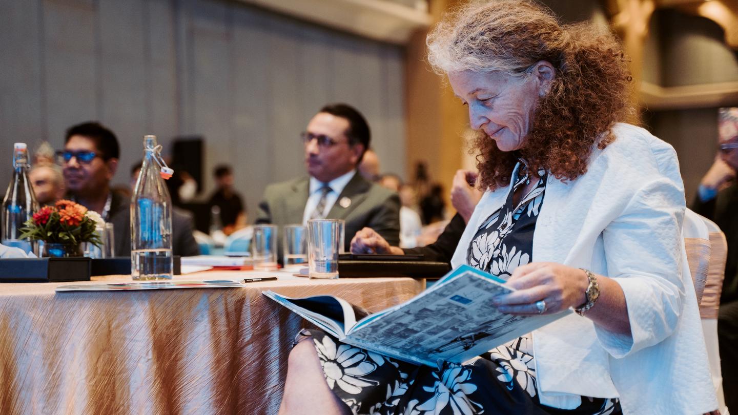 woman in white blazer reads a magazine at a conference table; other attendees chat in background