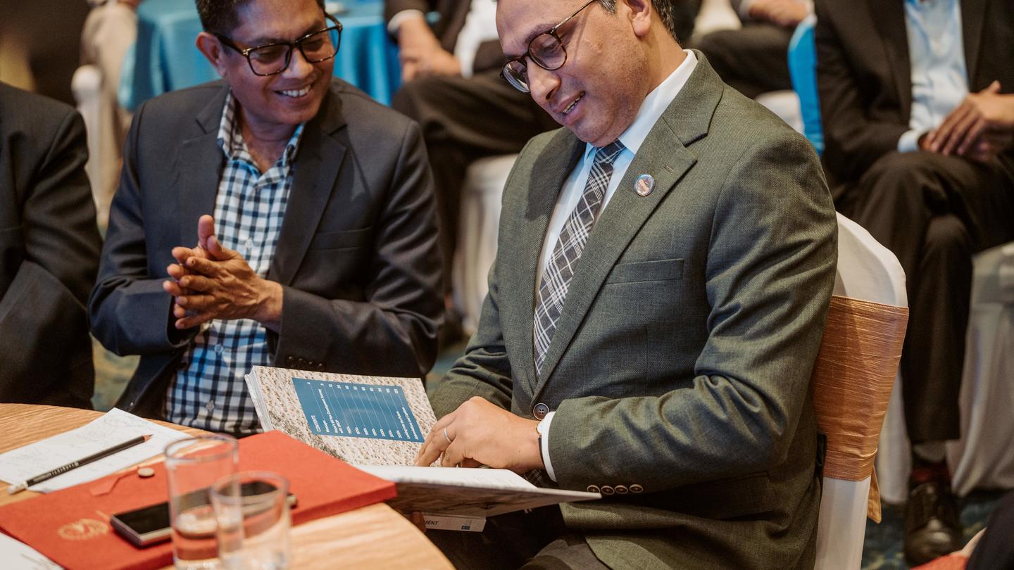 Photograph of two men at a conference table reviewing documents, with others in background.