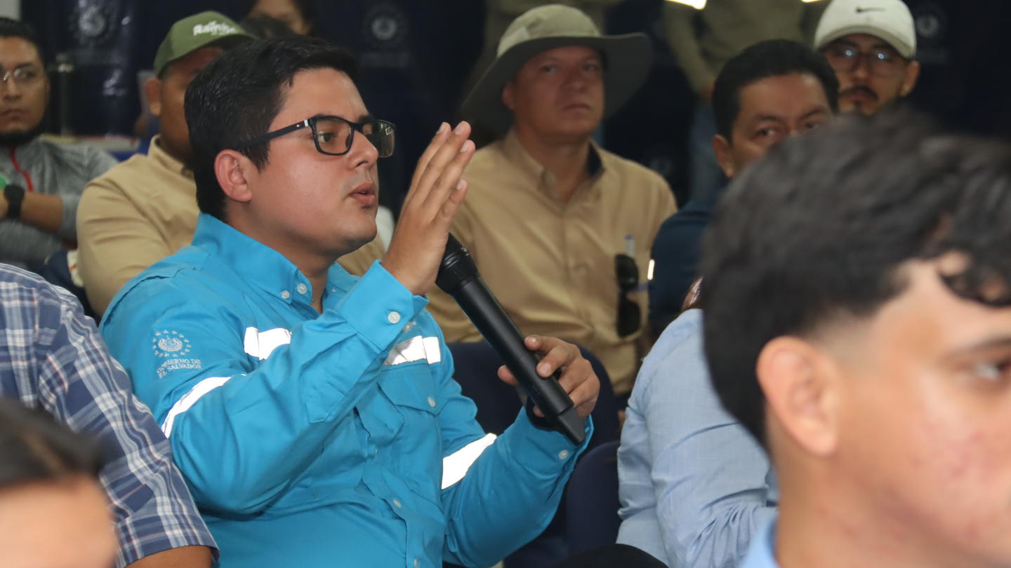 Audience member in blue shirt and glasses raises his hand at a conference.