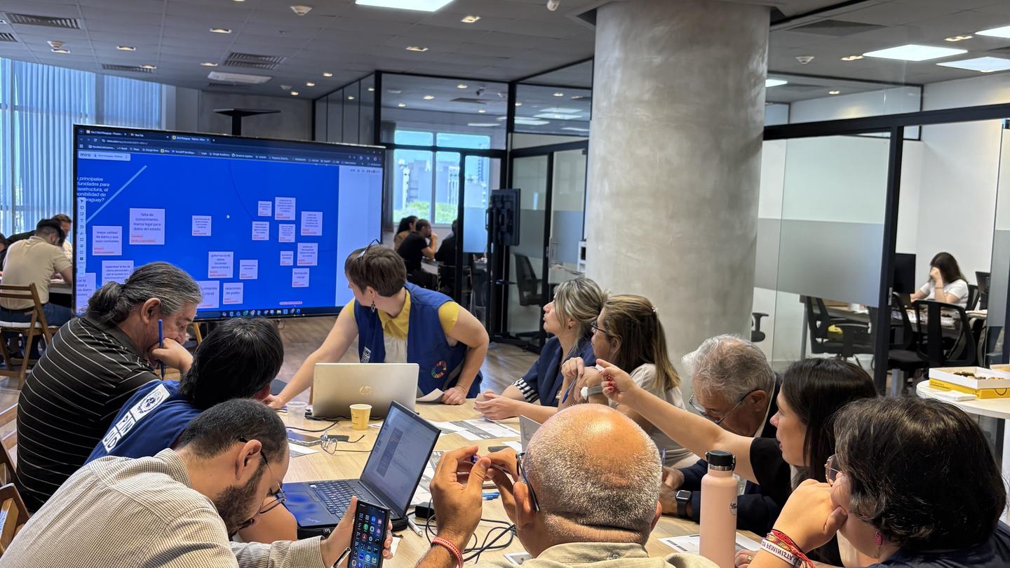 Photograph of a busy conference room with people around a long table and a blue screen in the background.