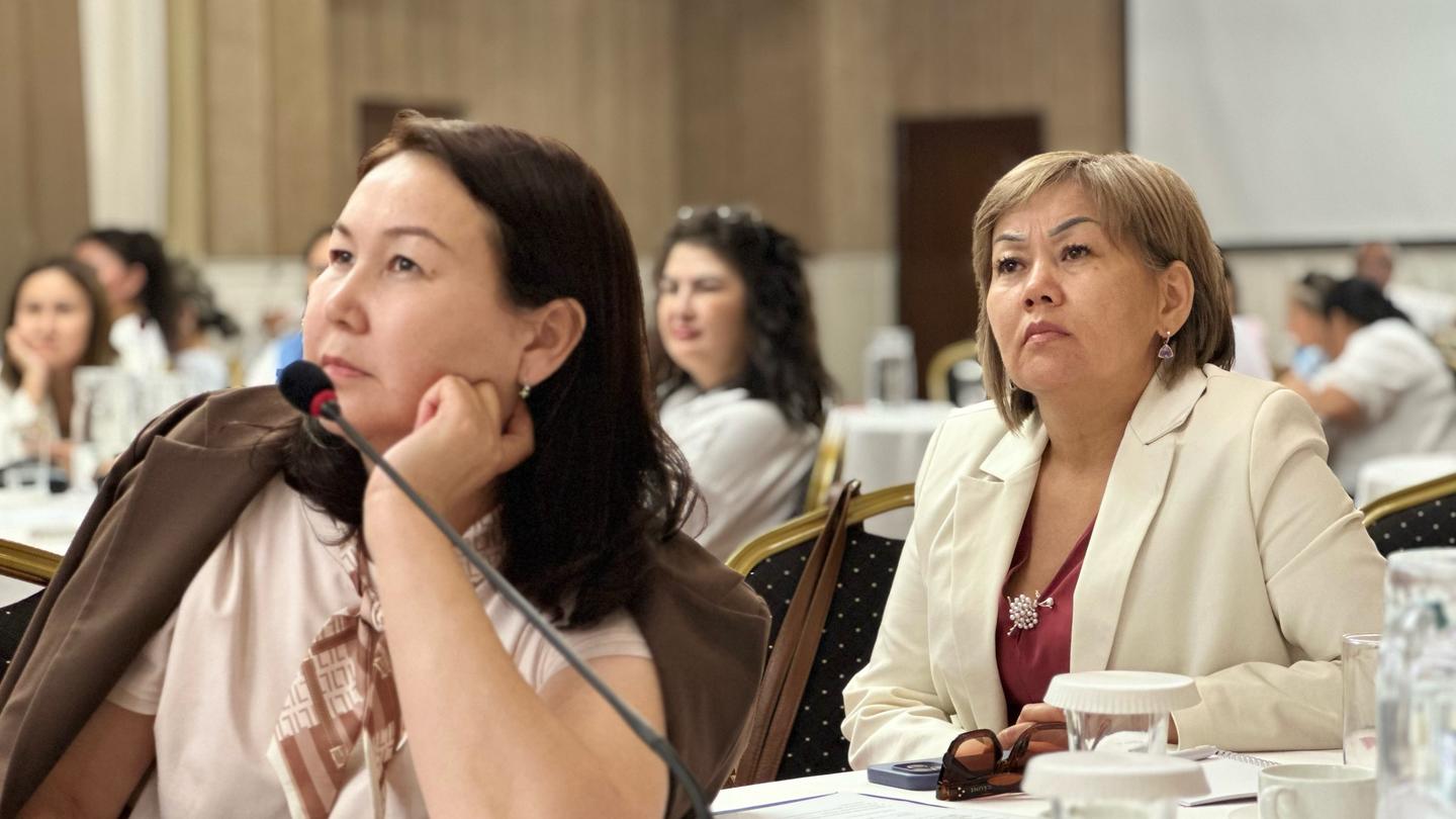 Photograph of two women at a conference table with a microphone; attendees seated in background.