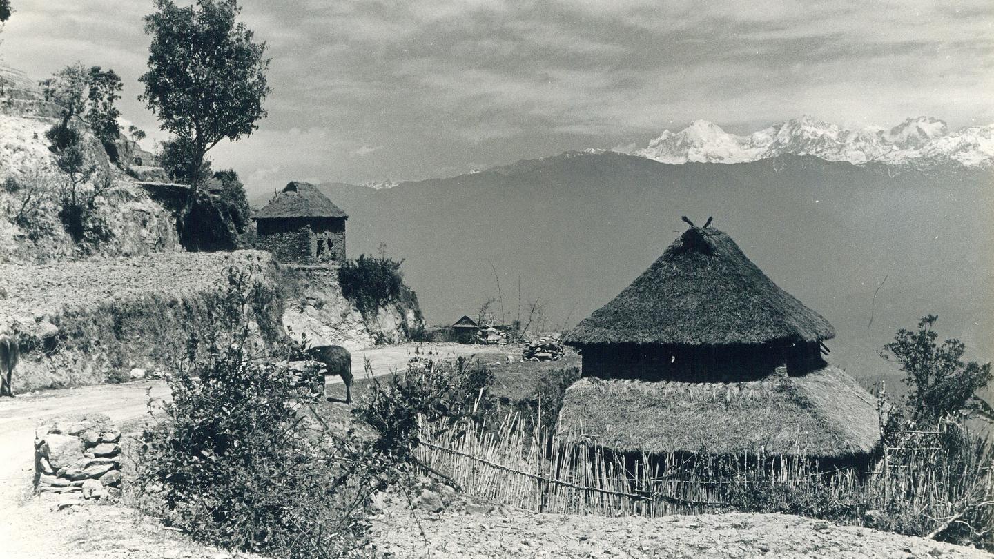 Black-and-white photo of hillside village with thatched round huts, stone walls, and distant mountains.