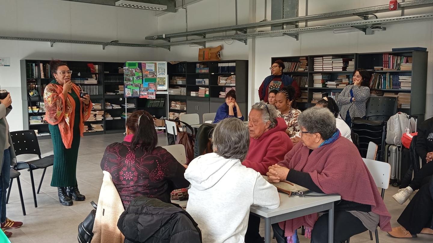 People seated around tables in a classroom workshop; a presenter at the front, shelves in back.