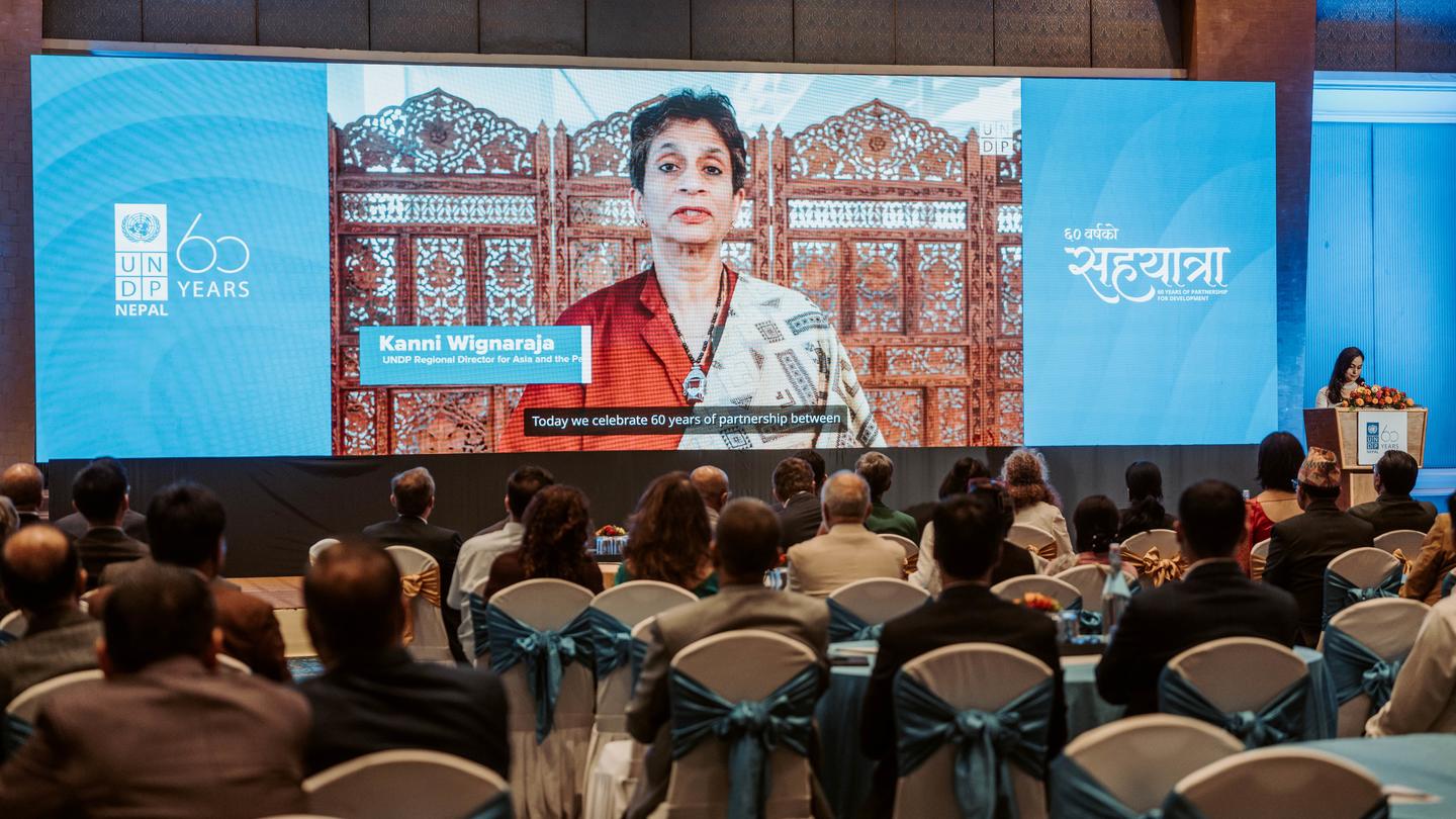 Photograph of a conference hall with audience watching a speaker on a large screen.