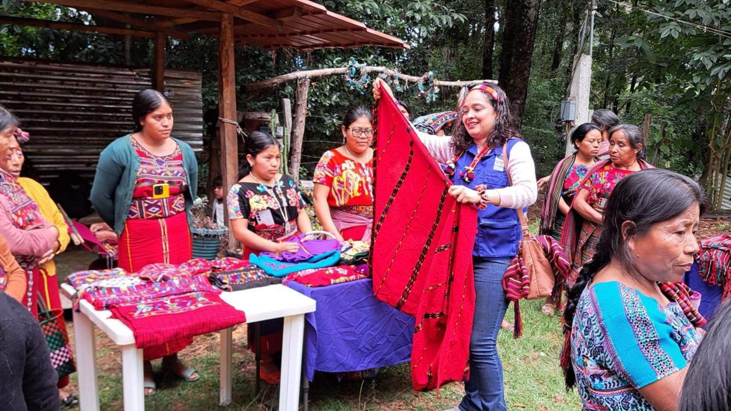 Outdoor community gathering around a red-draped table under a canopy; people wear colorful clothes.