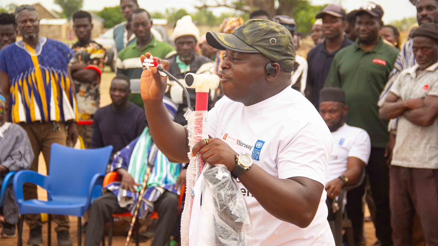 Man in white shirt and camouflage cap speaks into a microphone to an outdoor crowd.