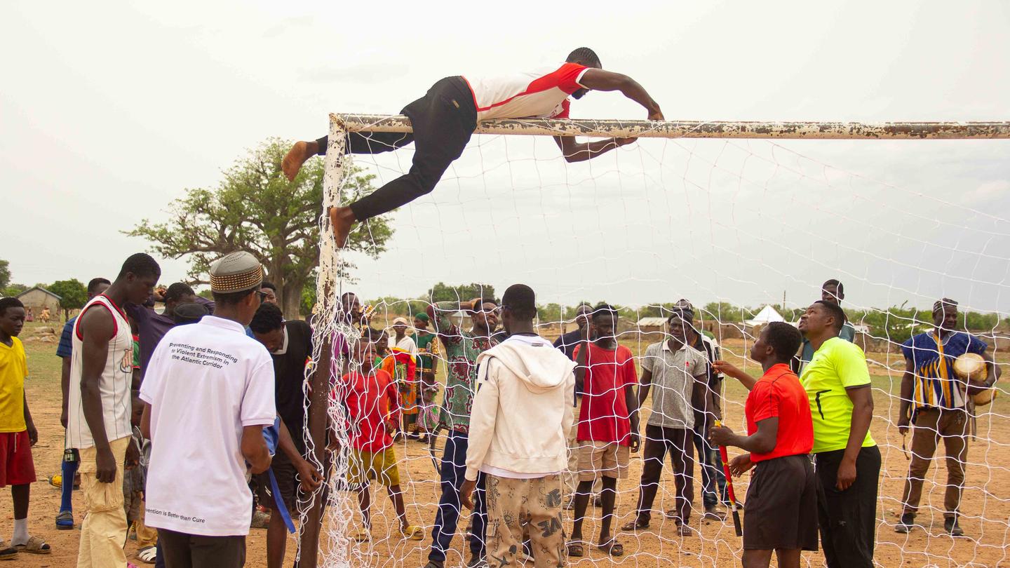 Man balancing on a long wooden pole held by supports, dusty outdoor crowd watches.
