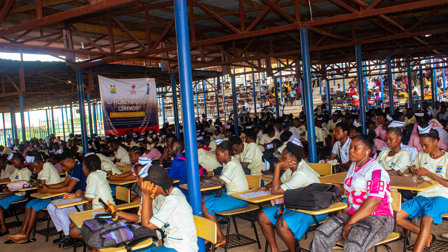 Photograph of a crowded classroom with students at long desks under a wooden ceiling and blue posts