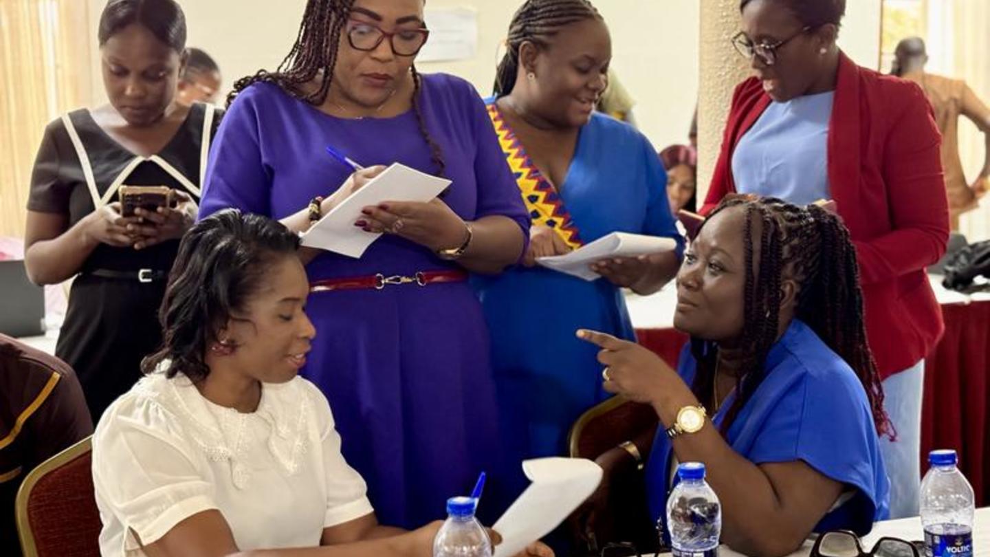 Diverse group of women at a workshop; some seated with notebooks, others stand with papers.