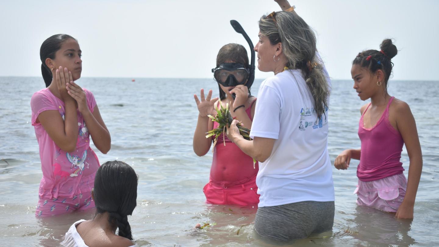 Group of people in pink swimsuits standing in shallow water at the beach; instructor raises hand.