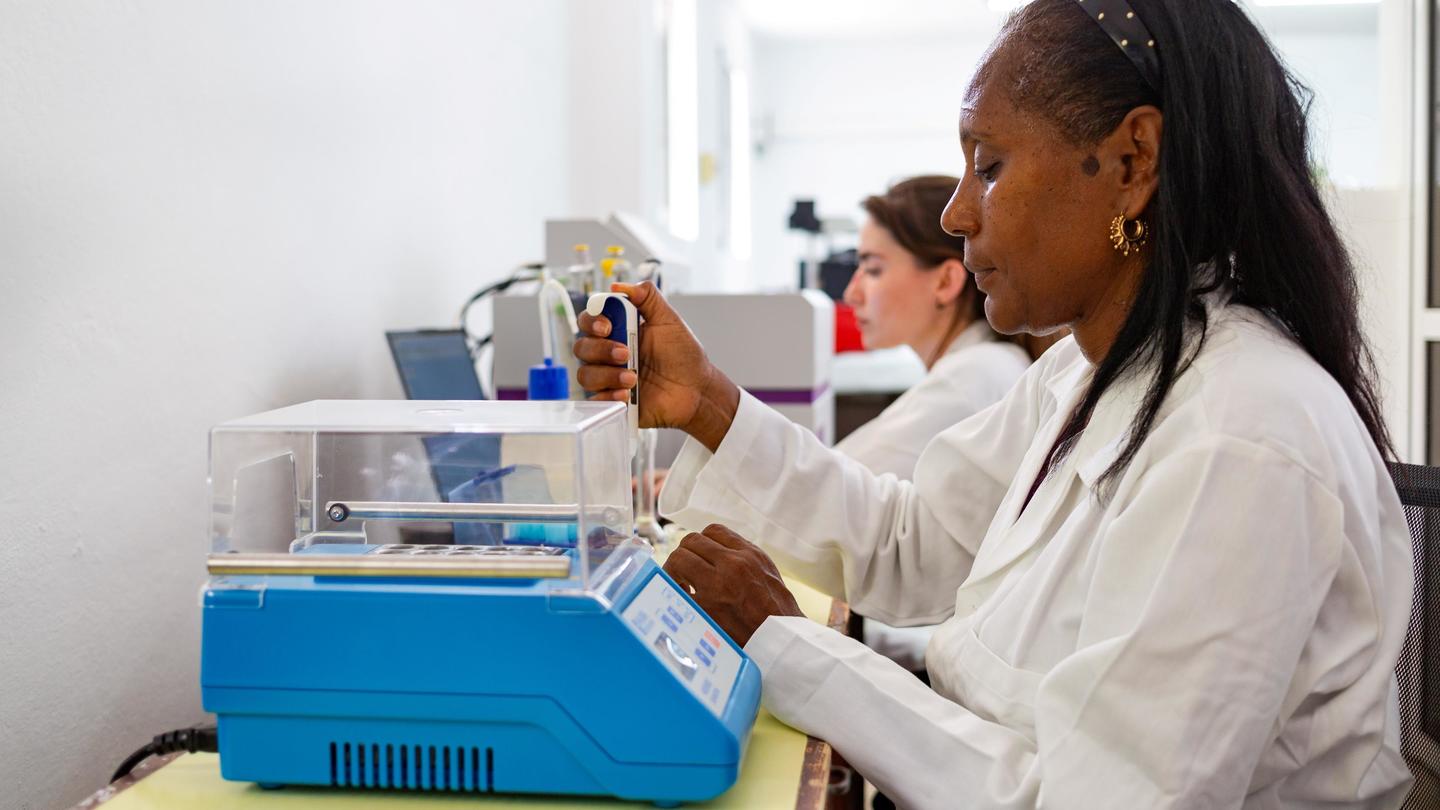 Scientists in white lab coats pipette into tubes beside a blue benchtop instrument.
