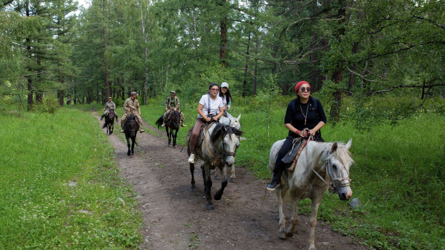 People on horseback ride along a dirt path through a lush green forest.
