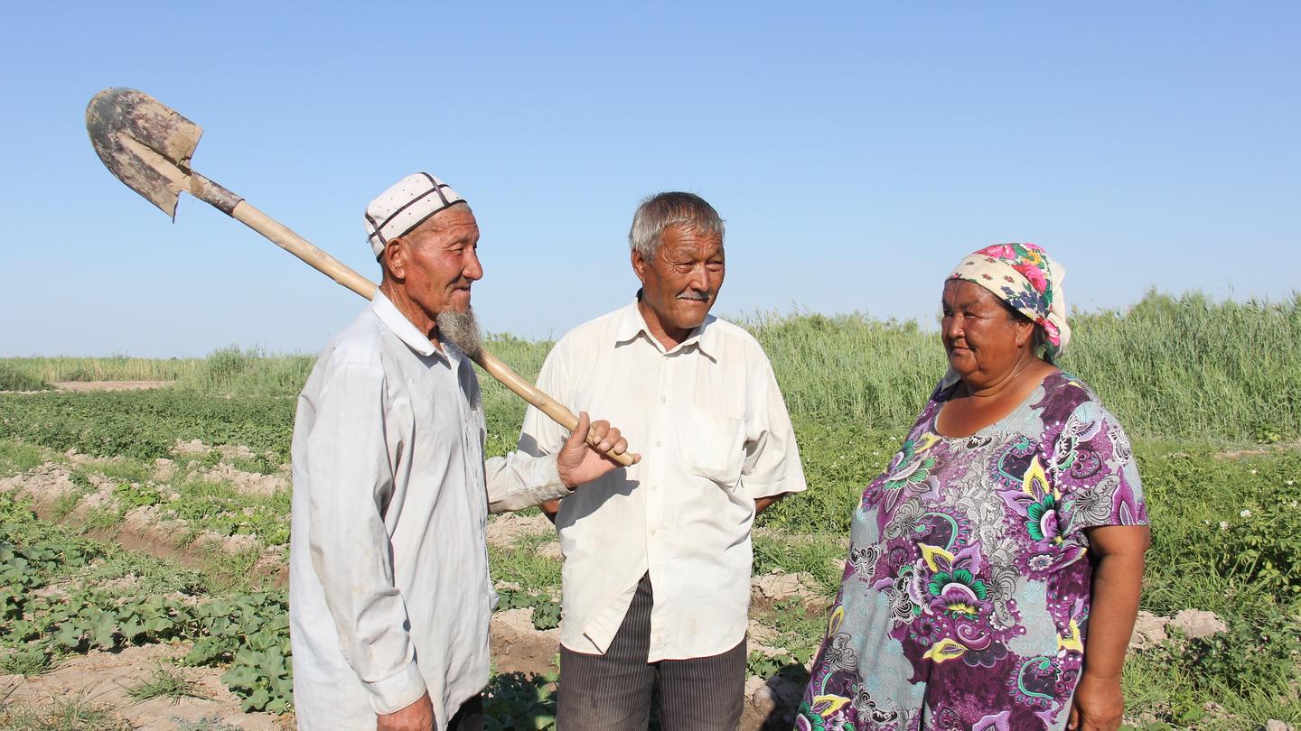 Three people stand in a sunlit field, conversing; one holds a long-handled tool with a curved blade.