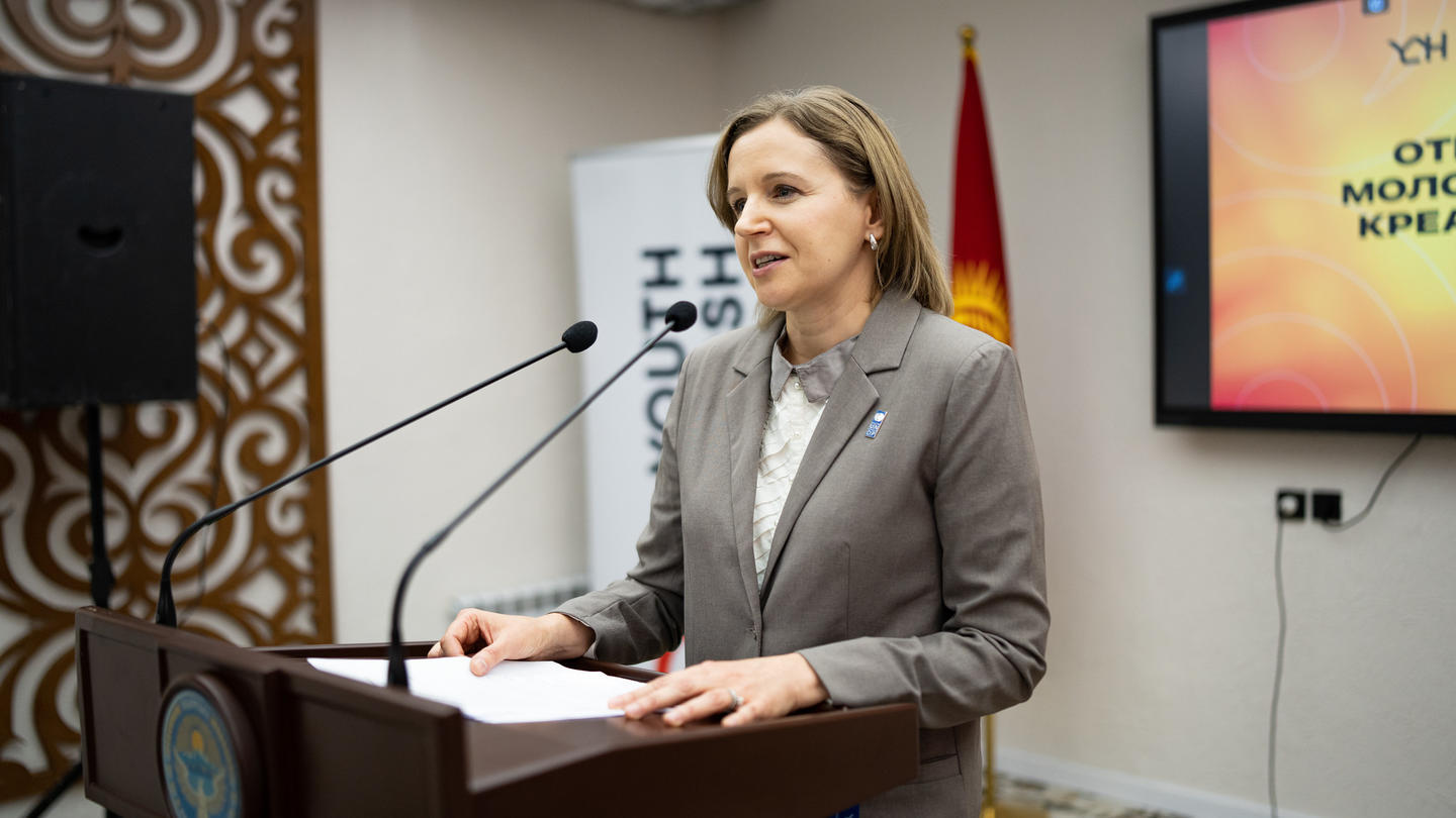 Photograph of a woman in a gray suit at a podium giving a speech in a conference room.