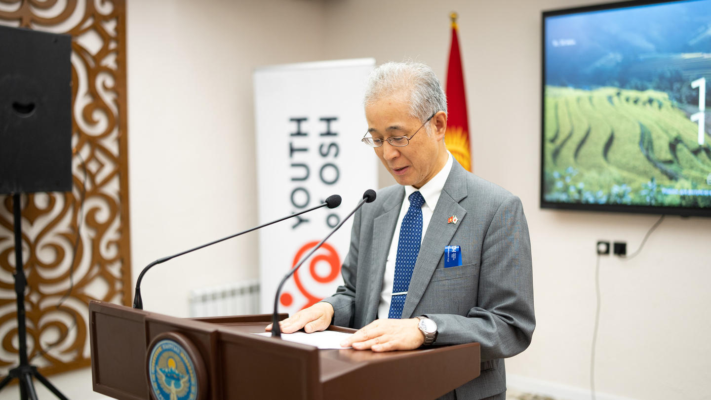 Senior man in a suit speaks at a podium with banners, flags, and a screen behind.