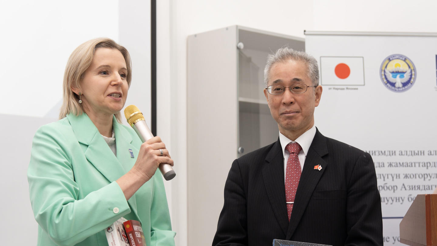 Photo: Woman in mint blazer speaks into microphone beside man in suit at a conference.