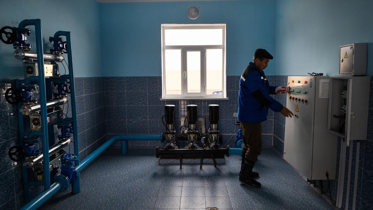 Technician in blue coveralls examines an electrical panel in a blue-walled workshop.