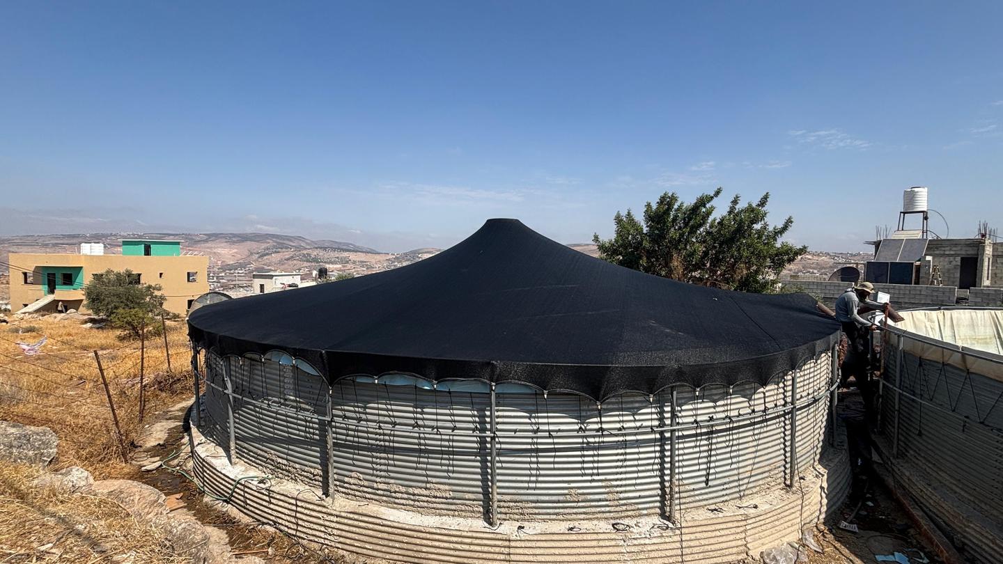 Round concrete water tank with black canopy, fenced, in a desert landscape with distant hills.