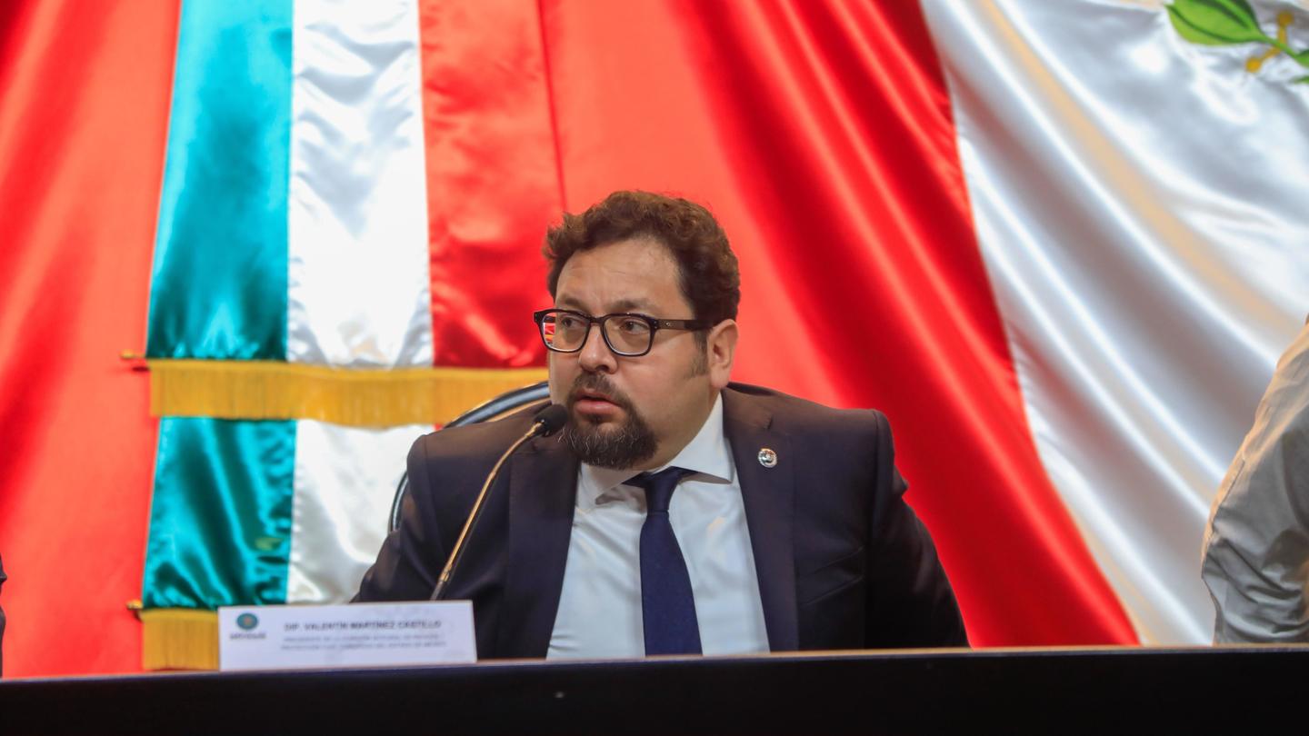 Man in suit and glasses speaks at a panel, with red, white, and blue flags behind.