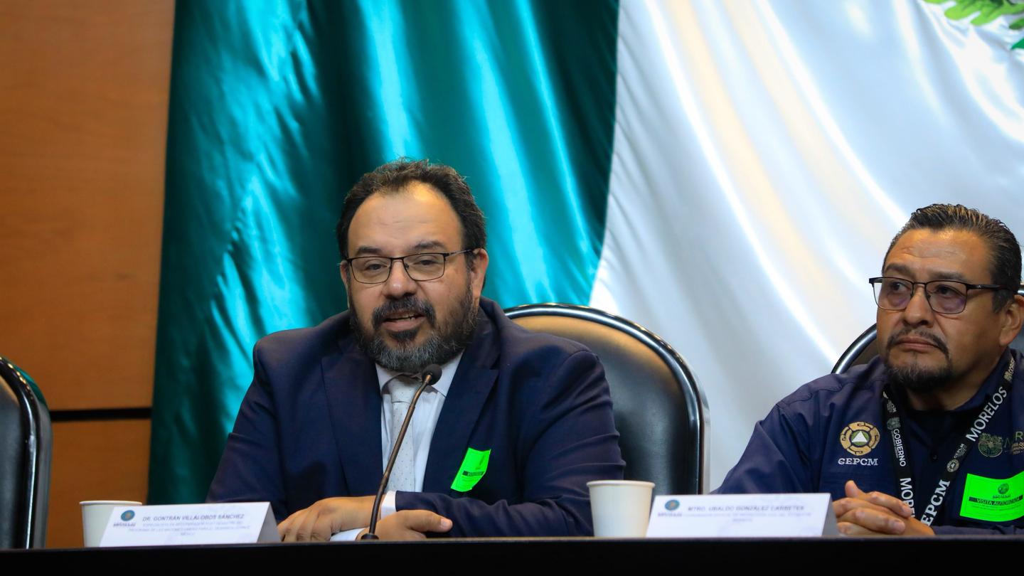 Two men with green badges sit at a conference panel, teal and white drapes behind.