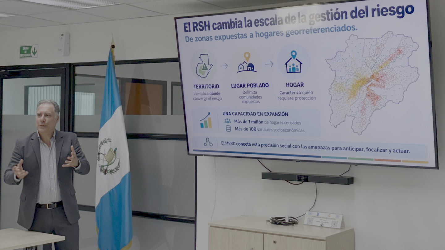 An older man in a blazer gestures beside a screen showing a map slide; a Guatemala flag is nearby.