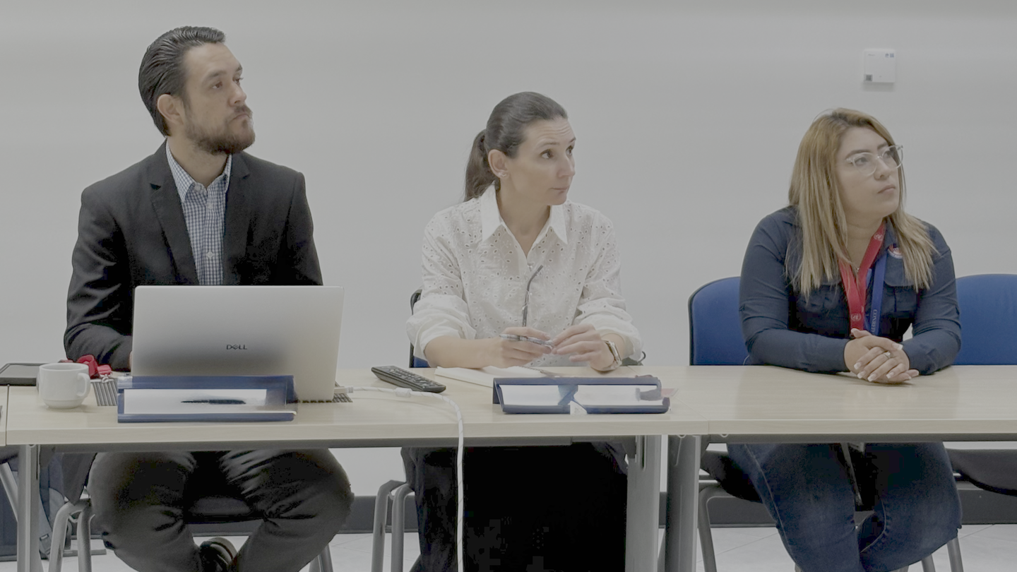 Three panelists seated at a conference table with laptops and notebooks, against a gray backdrop.