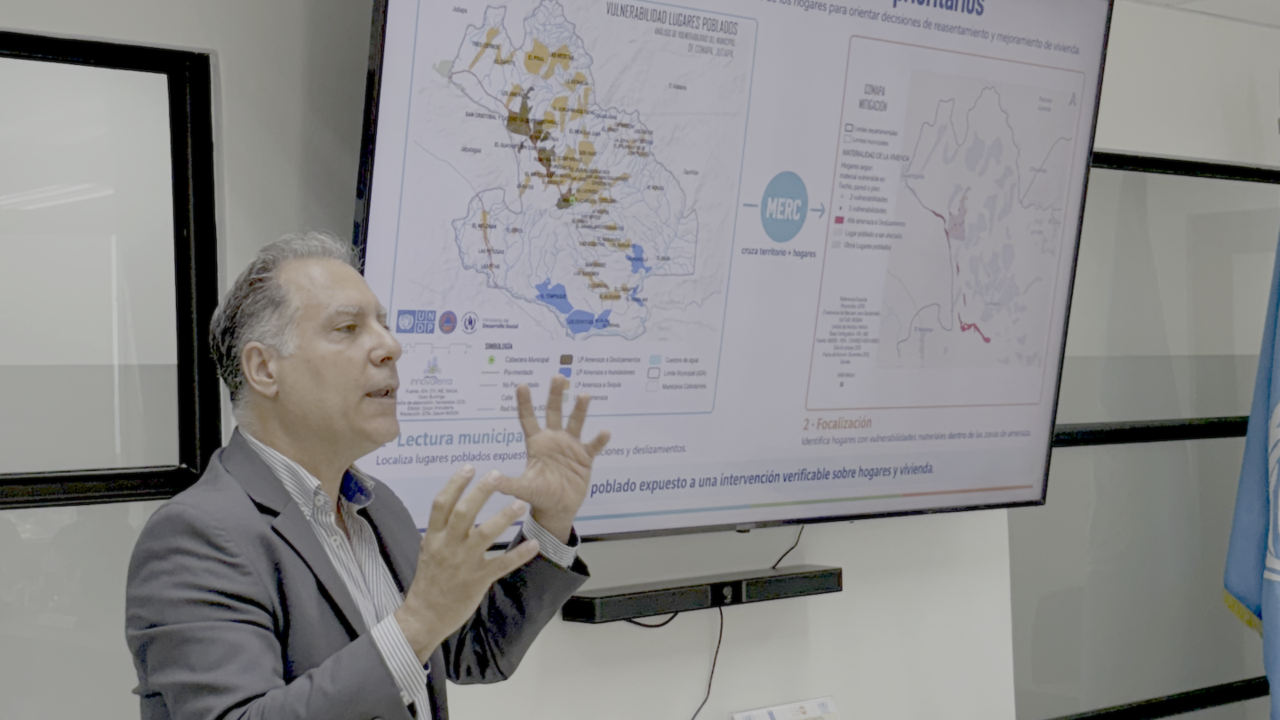 Photograph of a male presenter in a gray suit gesturing at a map on a large screen.